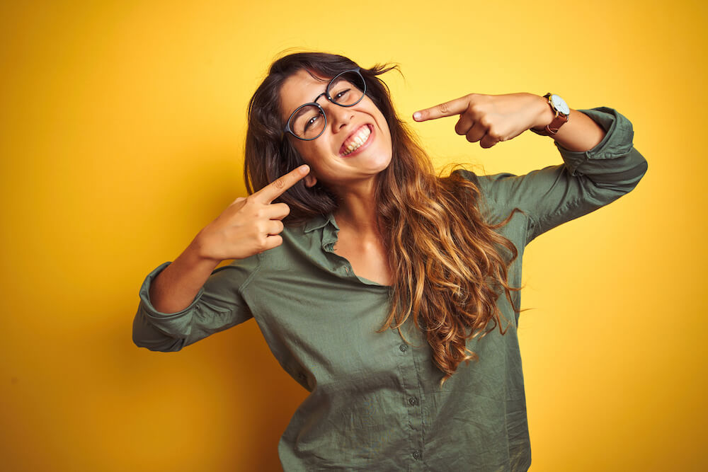 Young beautiful woman wearing green shirt and glasses over yelllow isolated background smiling cheerful showing and pointing with fingers teeth and mouth. Dental health concept.