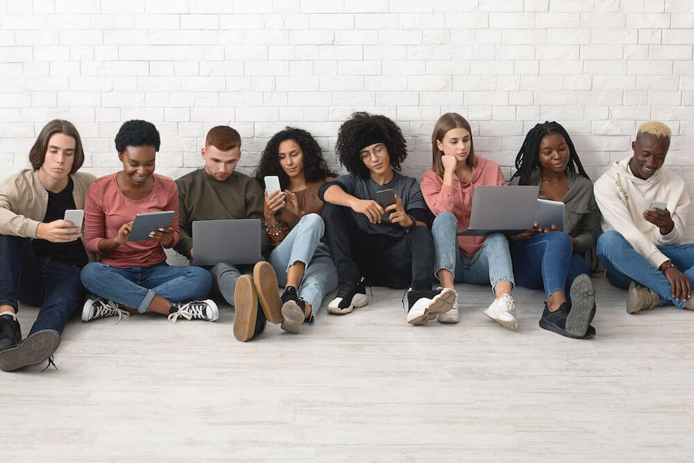 Gadget addiction in millennials concept. International group of positive young people sitting on floor next to white brick wall and using different gadgets notebooks, pads, mobile phones, panorama